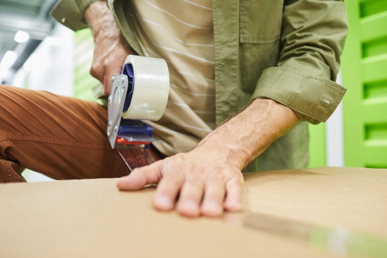 Close up of unrecognizable man sealing cardboard boxes with scotch tape gun while packing in self storage unit, copy space