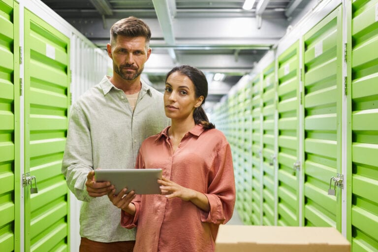 Waist up portrait of beautiful modem couple holding digital tablet while standing by self storage unit and looking at camera, copy space