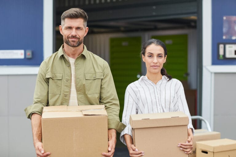 Waist up portrait of contemporary couple holding boxes and looking at camera while standing by self storage facility, copy space