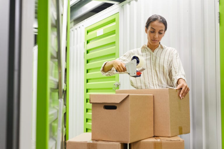 Portrait of young woman packing boxes with tape gun while standing by self storage unit, copy space